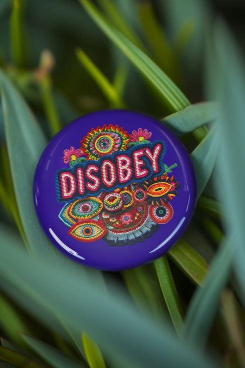 Stay Weird" black pinback button with pearl-effect lettering surrounded by folk art flowers in red and turquoise on a dark background.